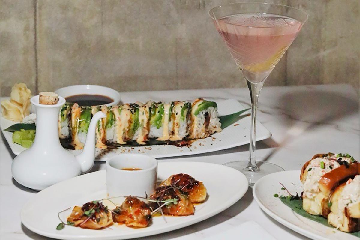 A table set with sushi rolls, dumplings, dipping sauces, a white soy sauce dispenser, and a pink cocktail in a martini glass, arranged on white plates against a neutral background.