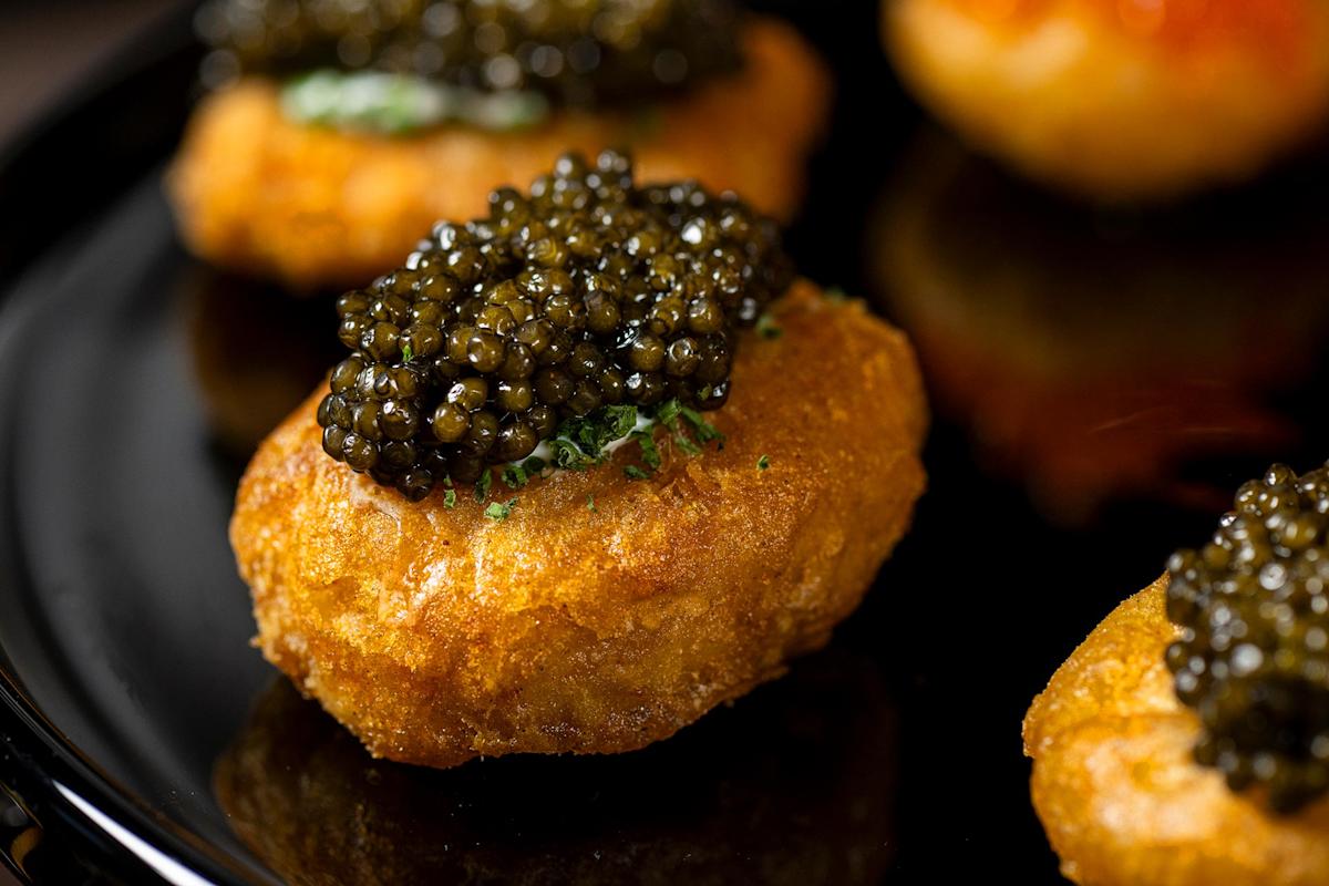 A close-up of a golden, crispy fried potato cake topped with a generous portion of glossy black caviar, served on a black plate.
