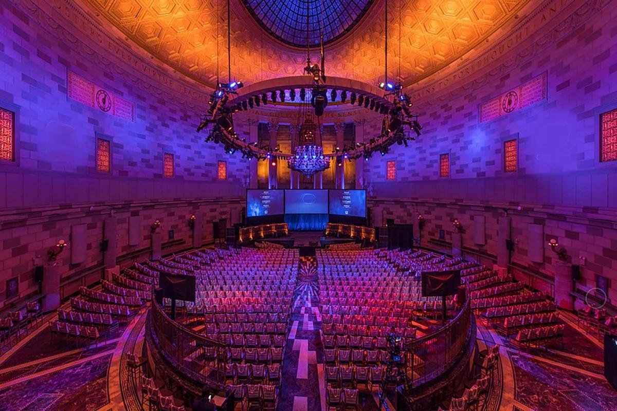 A large, ornate auditorium with rows of empty chairs, dramatic purple and orange lighting, a domed ceiling, and a central chandelier, set up for a stage event or conference.