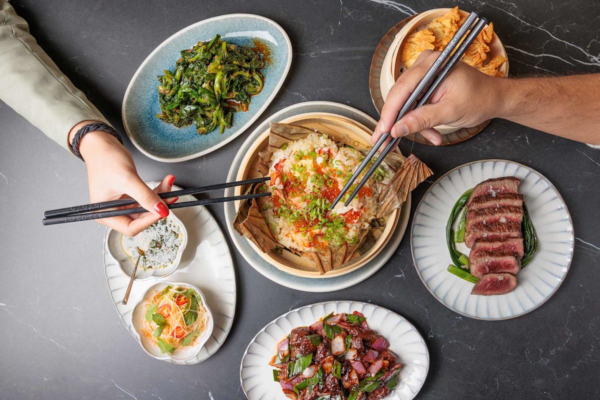 A table with various Asian dishes, including rice wrapped in leaves, sliced steak, sautéed greens, stir-fried beef, salad, and chips. Two people use chopsticks to take food from the central dish.