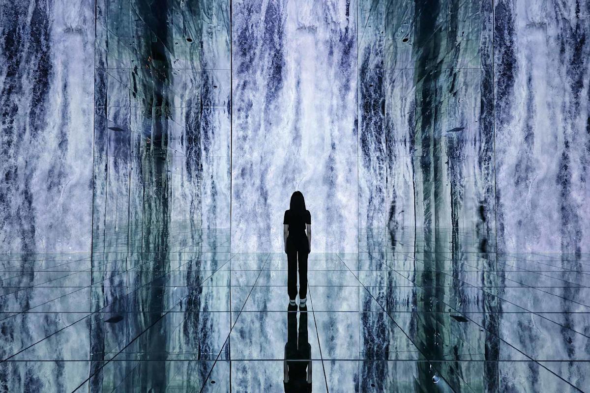 A silhouette of a person stands in front of a large illuminated waterfall display, surrounded by mirrored walls and floor reflecting the waterfall and their figure.