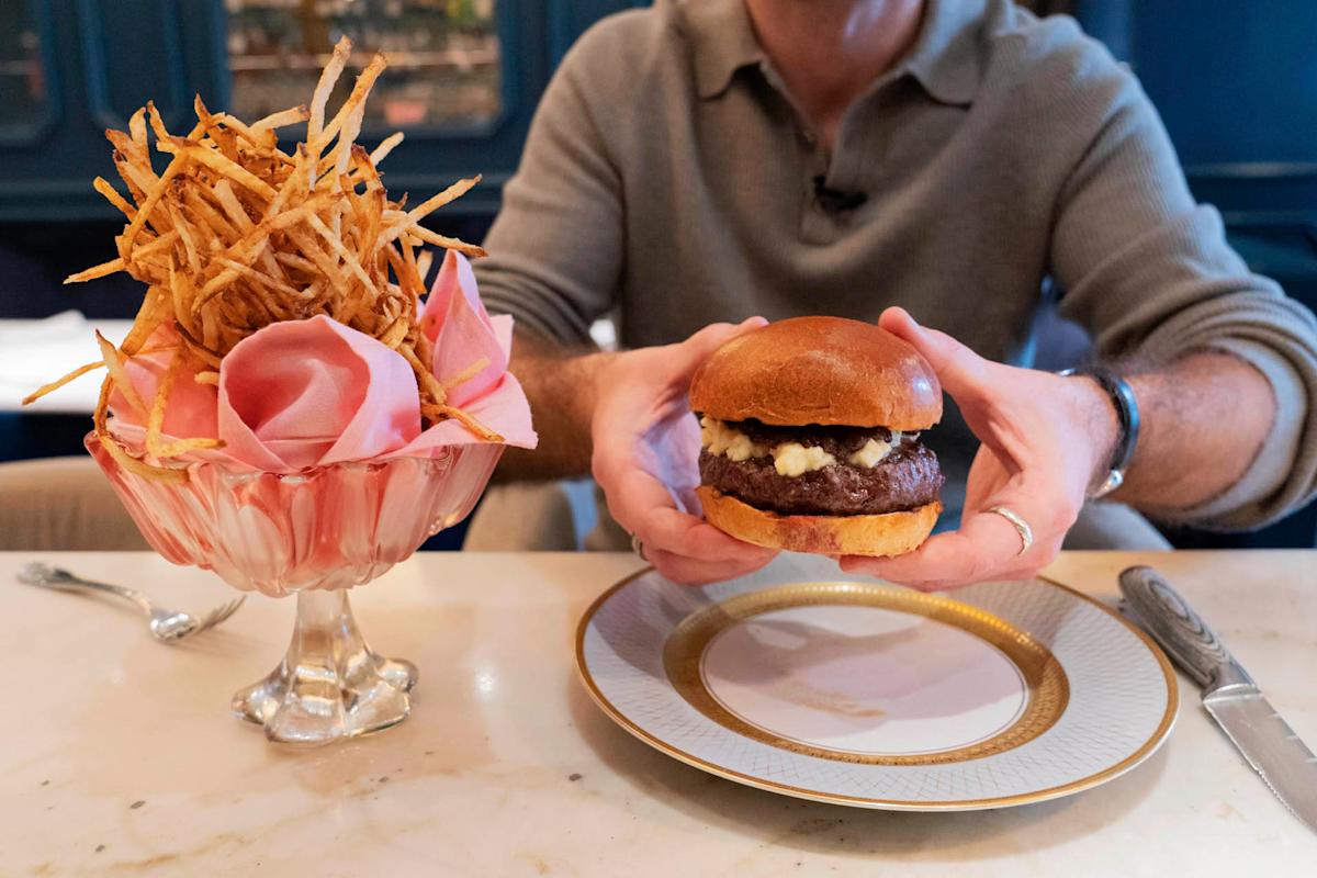 A person holds a gourmet hamburger on a decorative plate, with a tall glass bowl of thin, crispy fries beside it on a marble table. The person wears a beige long-sleeve shirt.