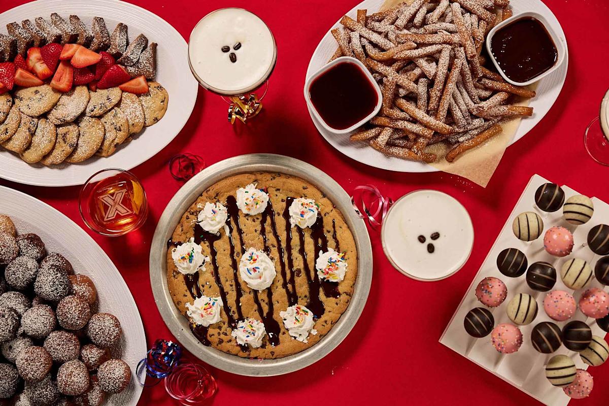 A variety of desserts on a red table, including a large cookie cake with whipped cream, churros with chocolate sauce, powdered donuts, chocolate truffles, and a plate of cookies with strawberries. Some drinks are also visible.