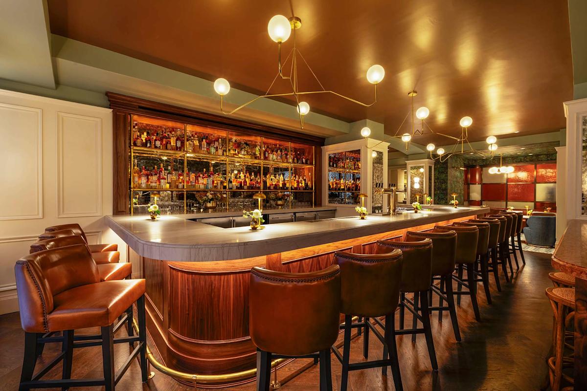 A stylish, modern bar with a marble countertop, brown leather barstools, and warm lighting. Bottles are displayed on wooden shelves behind the bar, and elegant pendant lights hang from the ceiling.