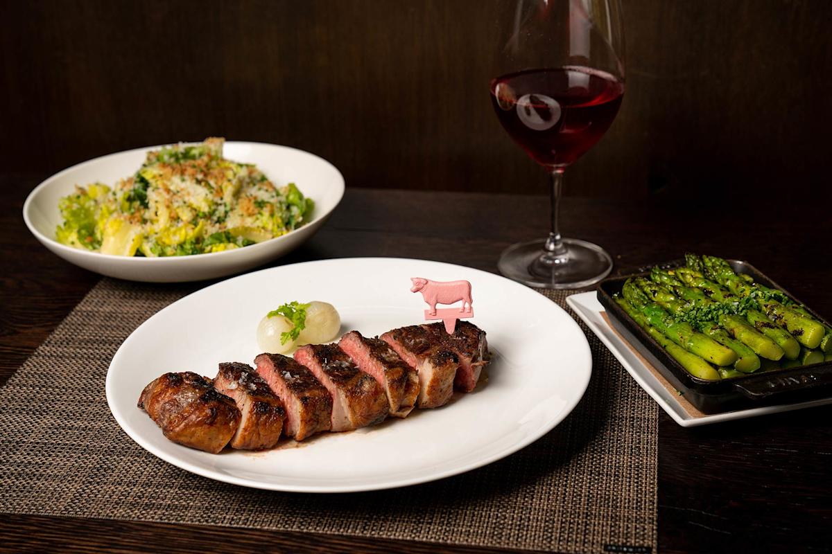 A plate of sliced steak with a small garnish and a pink cow marker, accompanied by a bowl of salad, a dish of asparagus, and a glass of red wine on a dark table.