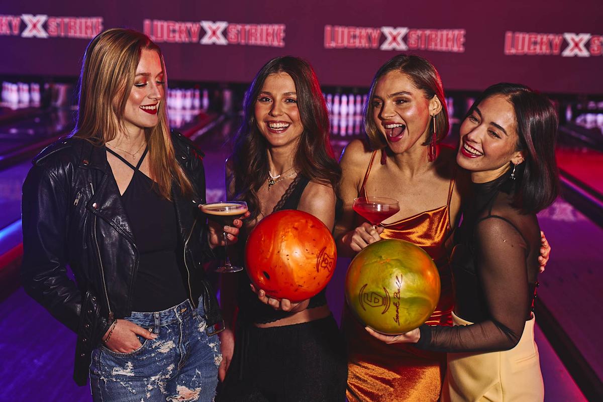 Four young women stand together at a bowling alley, smiling and holding drinks. Two of them hold brightly colored bowling balls. The background shows bowling lanes and illuminated signage reading "LUCKY STRIKE.