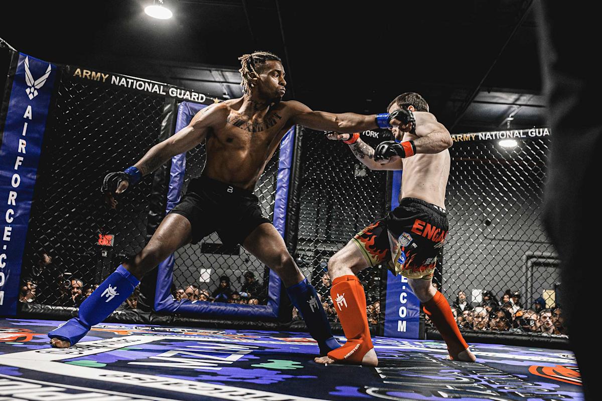 Two male MMA fighters compete in a cage match; one delivers a punch to his opponent’s face while both wear gloves, shin guards, and shorts. The audience watches from outside the octagonal cage.