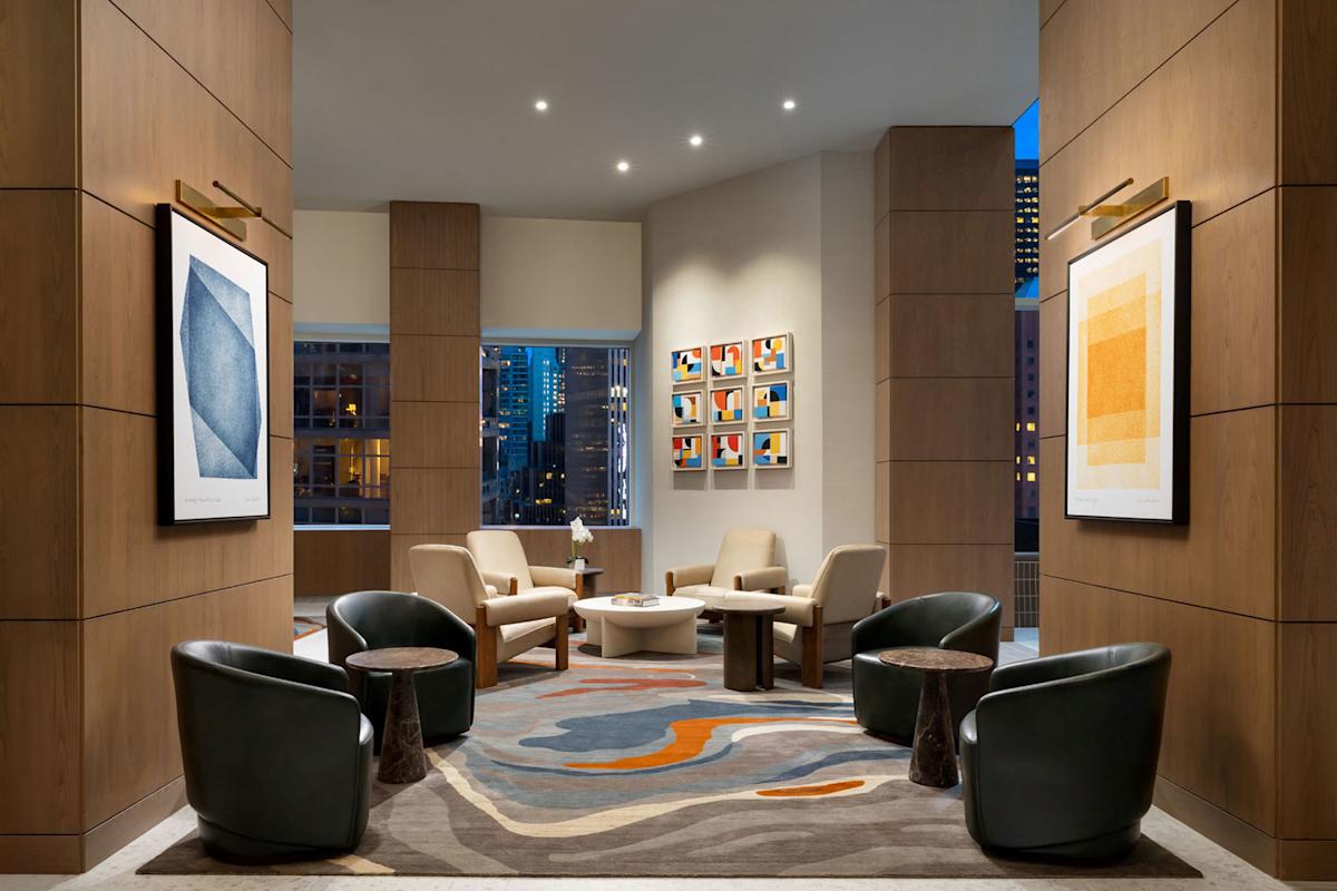 Modern lounge area with armchairs, round tables, abstract art on wood-paneled walls, a patterned rug, and large windows showing city buildings at dusk. Warm lighting creates an inviting atmosphere.