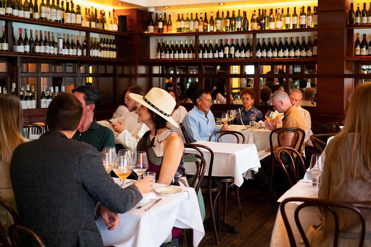 People dining in a cozy, elegant restaurant with wooden decor, white tablecloths, and shelves lined with wine bottles. Groups are engaged in conversation, and natural light fills the room.