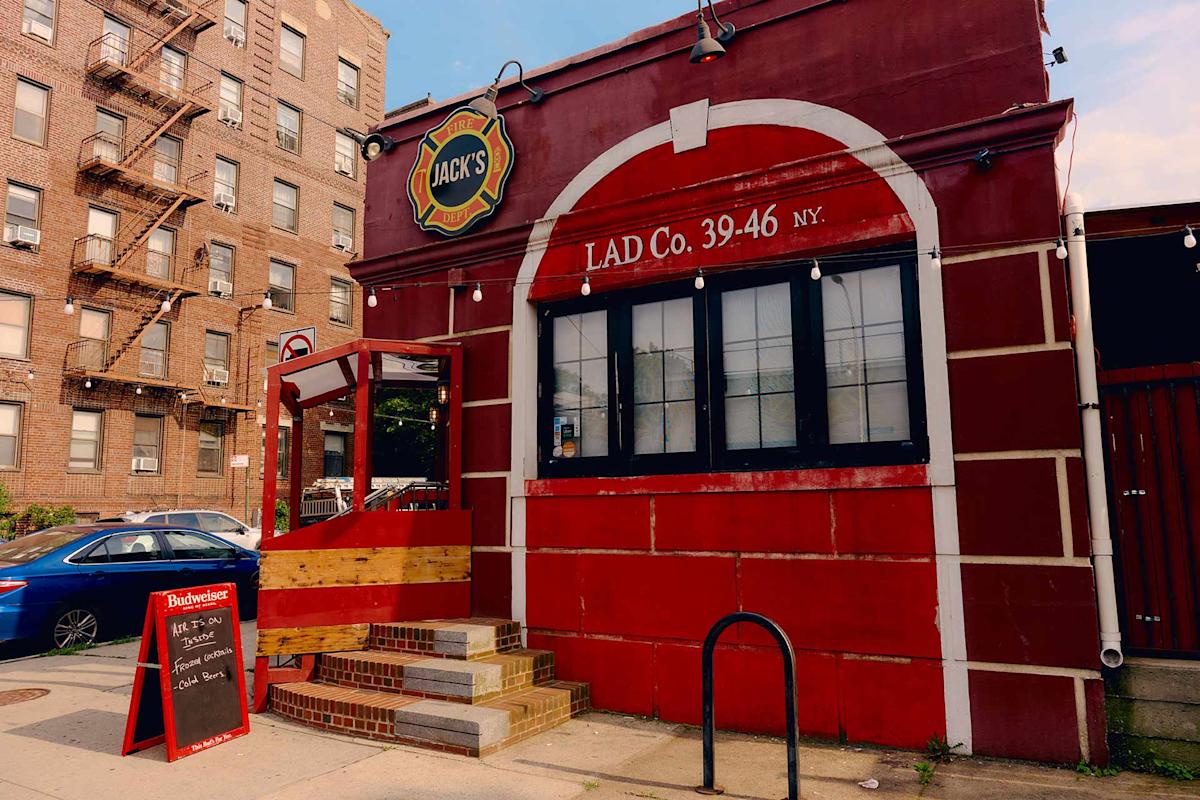A red brick restaurant called Jack’s with large front windows, a wooden entryway, and a sandwich board menu on the sidewalk. An apartment building and a blue car are visible in the background.