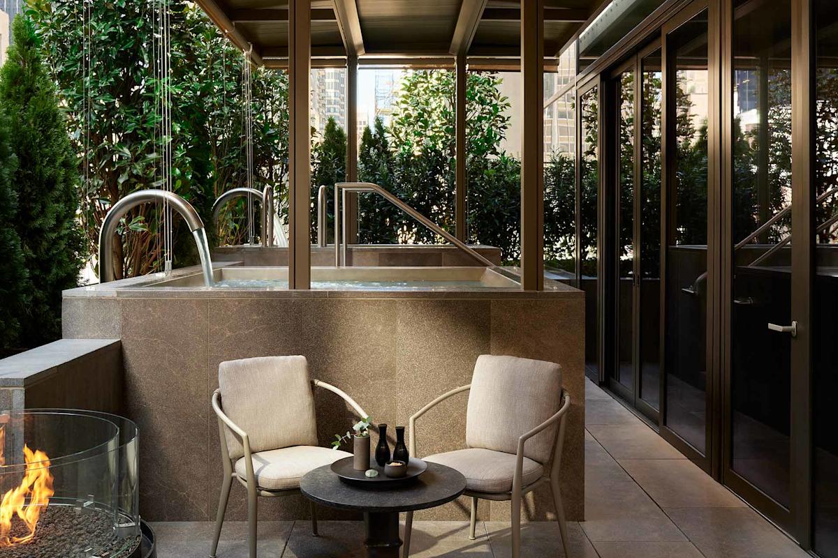 A private outdoor patio features two cushioned chairs and a small round table beside a modern fire pit, with a raised stone hot tub surrounded by greenery in the background. Glass doors line one side of the patio.