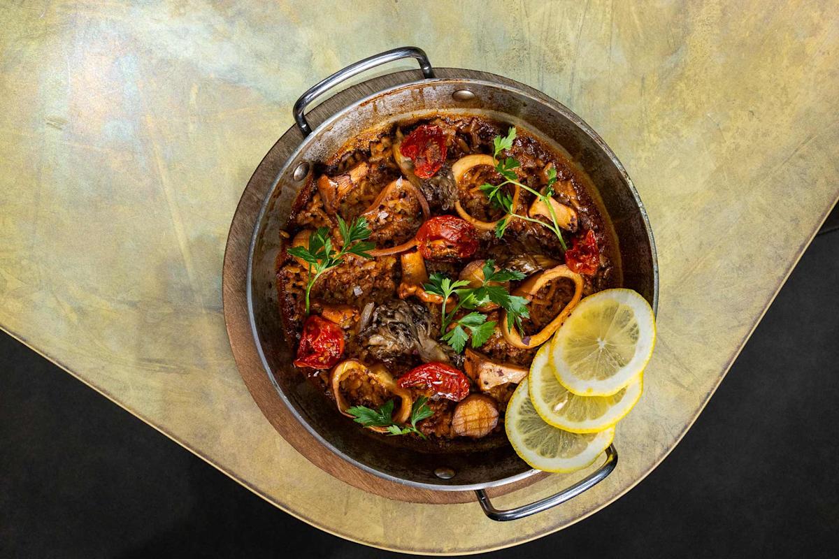 A round metal pan filled with a seafood stew containing shrimp, squid, tomatoes, and herbs, garnished with fresh parsley and three lemon slices, placed on a gold-colored table.