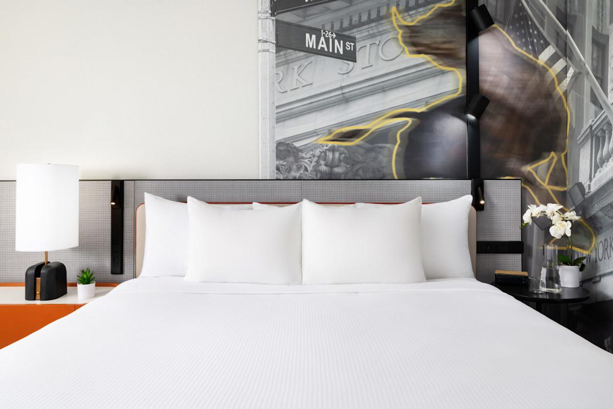 A neatly made hotel bed with white linens and pillows, flanked by modern nightstands, a lamp, and flowers. The headboard features a black-and-white mural with "Main St" and a blurred image of a bull.