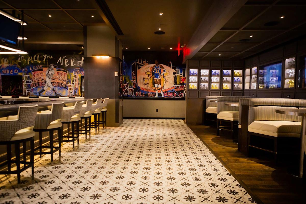 A stylish restaurant interior with patterned tile floors, cushioned chairs, a bar area on the left, wall art with bold colors in the center, and illuminated photo displays on the right. The space is warmly lit and inviting.