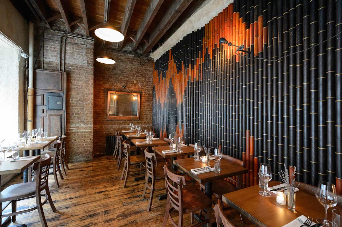 Cozy restaurant interior with wooden tables and chairs, exposed brick walls, wooden ceiling beams, and a unique black and orange textured wall design. The space is softly lit with warm hanging lights and candles on the tables.