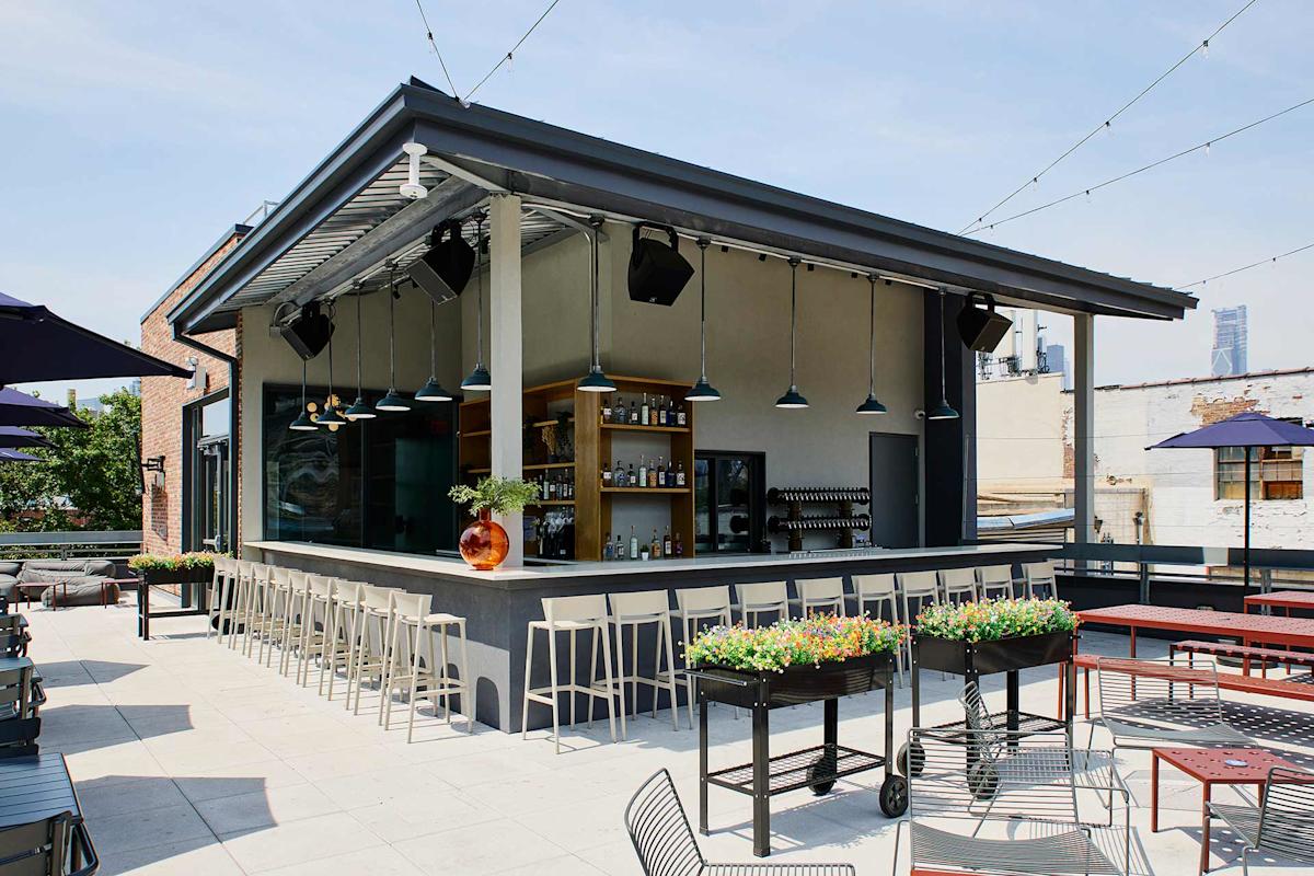 Modern rooftop bar with a central counter surrounded by barstools, hanging lights above, shelves stocked with bottles, planter boxes with flowers, string lights overhead, and outdoor tables and chairs under a clear sky.