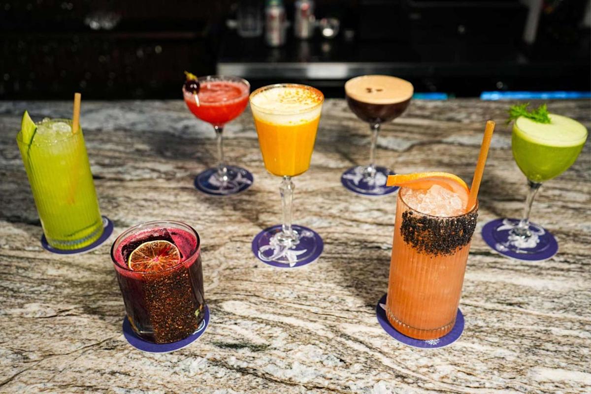 A variety of colorful cocktails and mocktails in different glasses are arranged on a marble countertop, each on a blue coaster. The drinks include green, yellow, red, orange, and purple beverages.