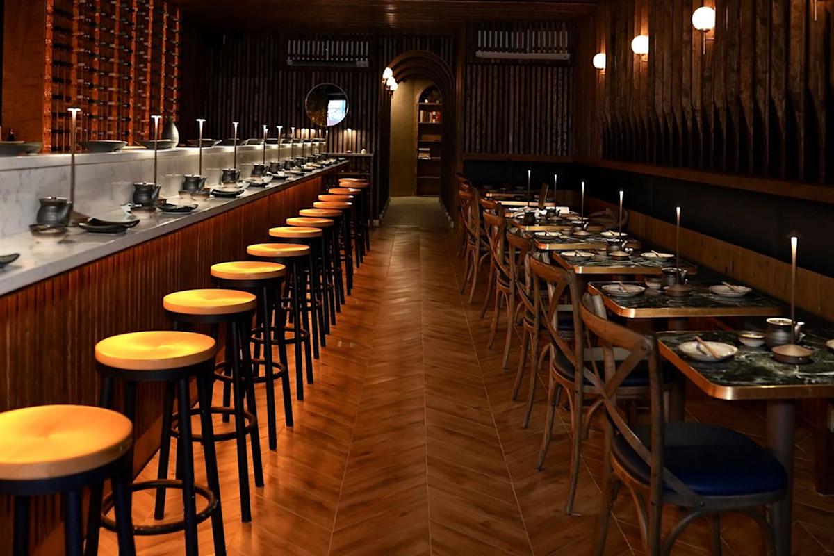 A warmly lit restaurant with a row of wooden stools at a marble counter on the left and wooden tables with chairs set for dining on the right, featuring candles and modern decor.