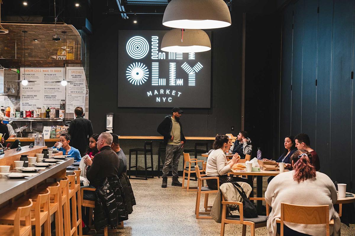 A modern indoor food market with people dining at tables and a bar, featuring a large illuminated sign reading “Ollie Market NYC” on the back wall. One person stands near the sign while others are seated and eating.