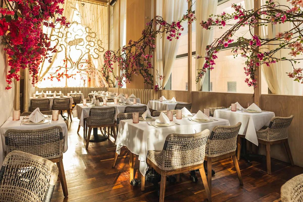 Sunlit restaurant with elegant white tablecloths, wicker chairs, and napkins neatly folded on tables. Pink blossom branches decorate the space, and large windows with sheer curtains let in natural light.