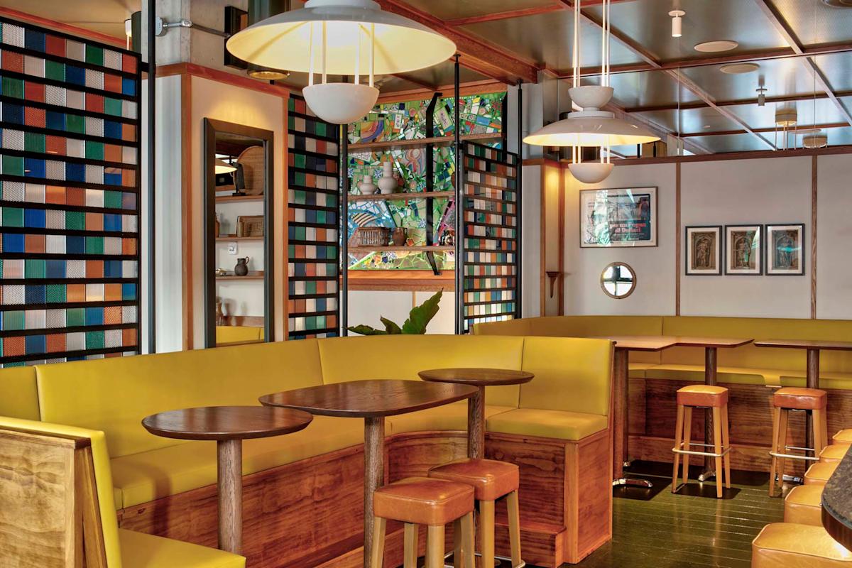 A stylish café interior with yellow cushioned benches, wooden tables and stools, colorful glass partitions, and framed artwork on the walls. Pendant lights hang from the ceiling, creating a warm and inviting atmosphere.