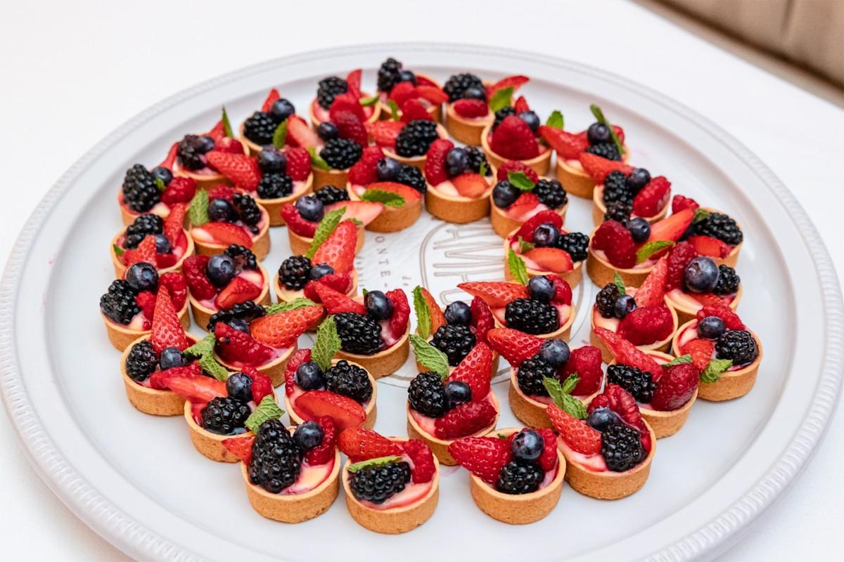 A white platter filled with small fruit tarts, each topped with strawberries, blackberries, blueberries, and mint leaves, arranged in concentric circles on a white tablecloth.