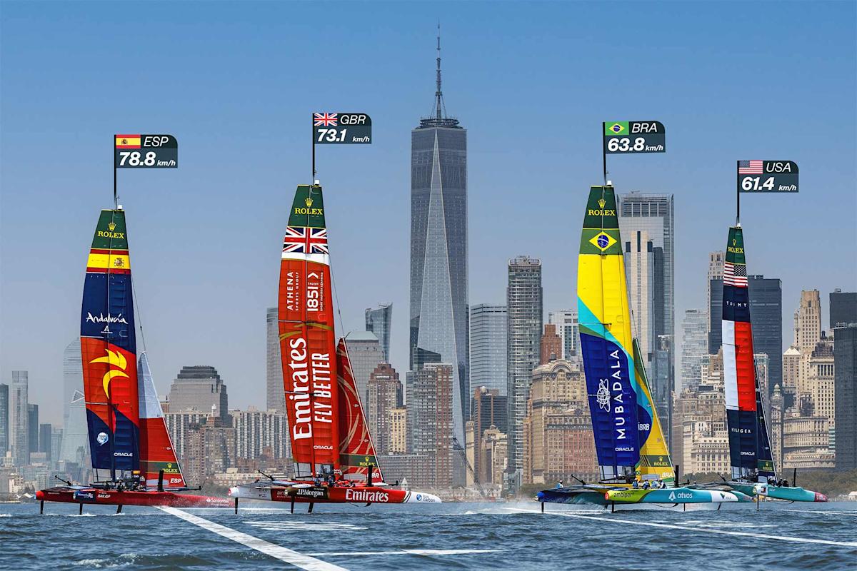 Four colorful sailboats from Spain, Great Britain, Brazil, and the USA race on the water with the New York City skyline and One World Trade Center in the background. Speed indicators appear above each boat.