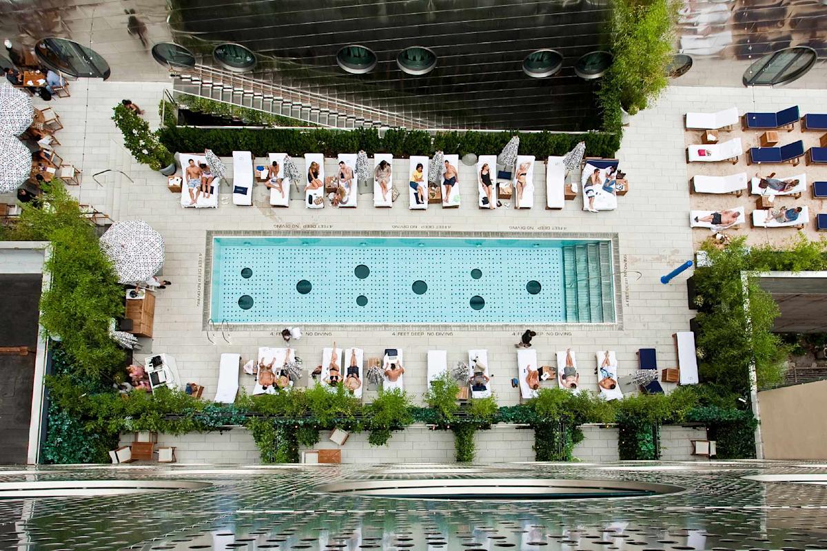 Aerial view of a modern outdoor swimming pool surrounded by people lounging on white chairs, with green plants and umbrellas lining the pool area.