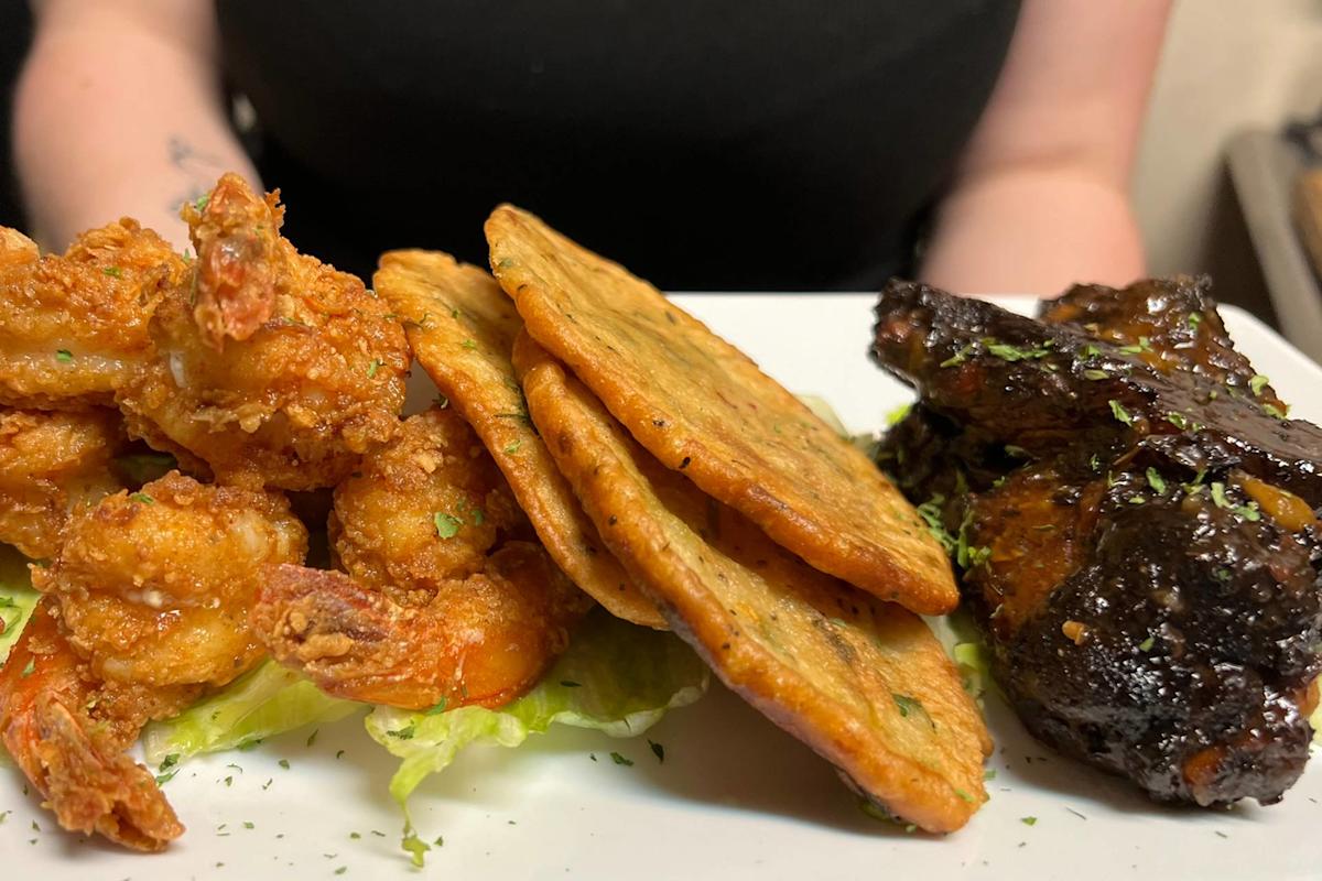 A plate with fried shrimp on lettuce, three golden-brown savory patties, and glazed, herb-topped chicken wings, all arranged neatly in front of a person wearing a black shirt.
