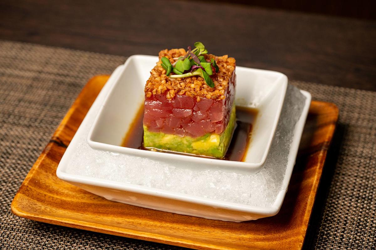 A square stack of diced raw tuna on top of avocado, topped with crispy bits and microgreens, sits in soy sauce on a white dish over ice, presented on a wooden tray.