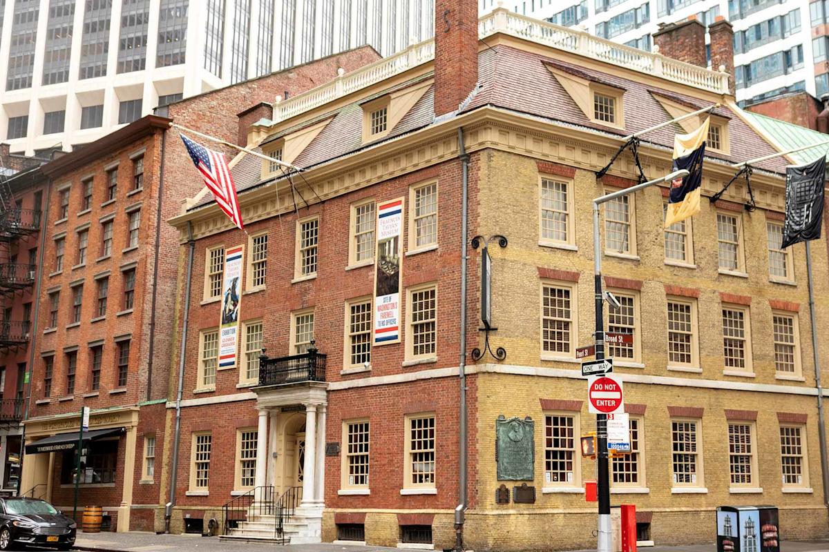 A historic brick building with cream trim and several flags displayed, located on a city street corner with modern skyscrapers in the background. A red "Do Not Enter" sign is visible at the intersection.