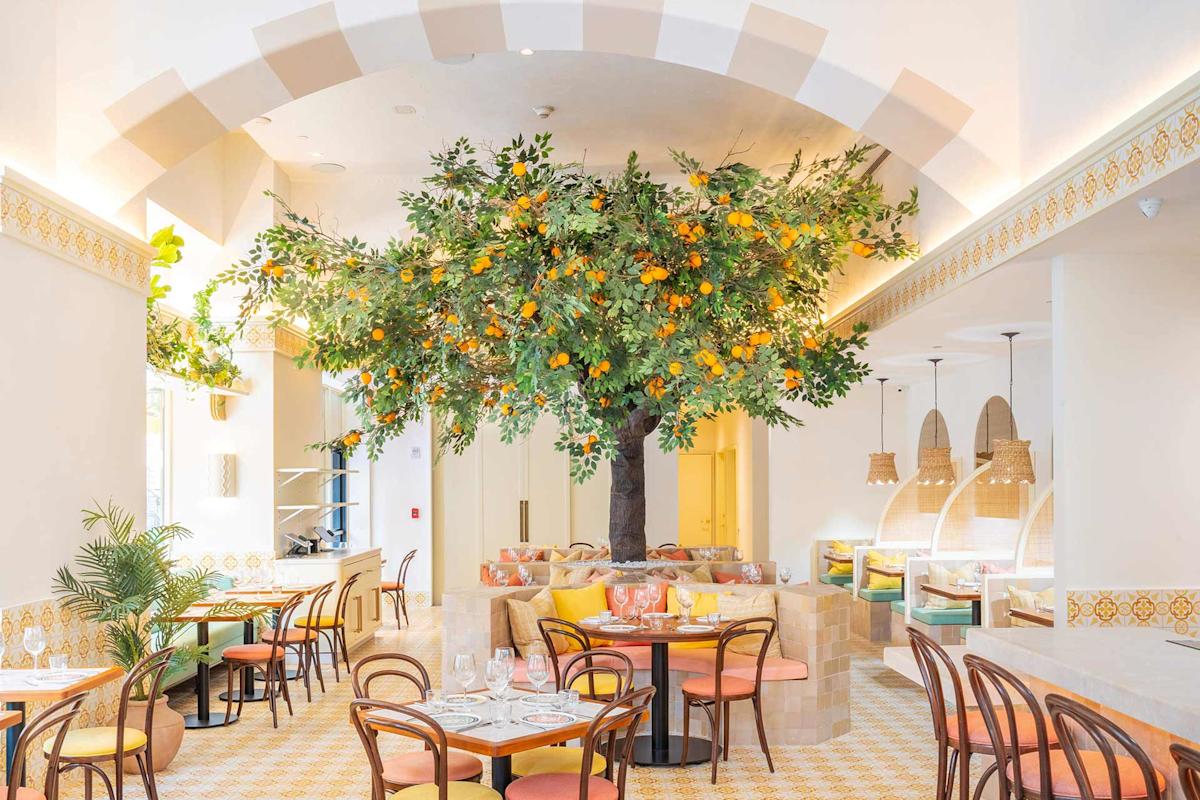 A bright, airy restaurant interior features a large artificial orange tree as its centerpiece, surrounded by tables and chairs, woven basket light fixtures, and pastel-colored decor with Mediterranean-inspired patterns.