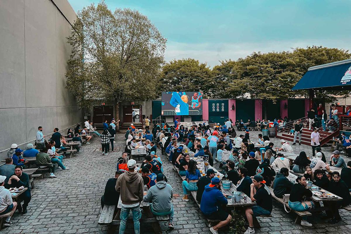 A large crowd of people sits at picnic tables in an outdoor courtyard, watching a sports game on a big screen. Trees and buildings surround the area, and the atmosphere is lively and social.