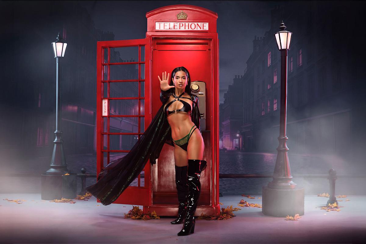 A woman in a black and green outfit poses with one hand raised in front of an open red British telephone booth on a foggy, dimly lit street at night, with scattered autumn leaves and old-fashioned street lamps.