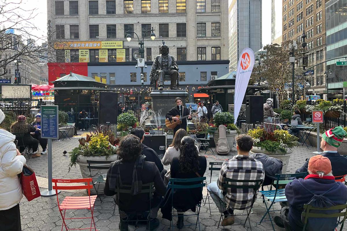 A musician plays guitar and sings in an outdoor plaza, performing to an audience seated on colorful chairs. Tall buildings and a large statue are visible in the background.
