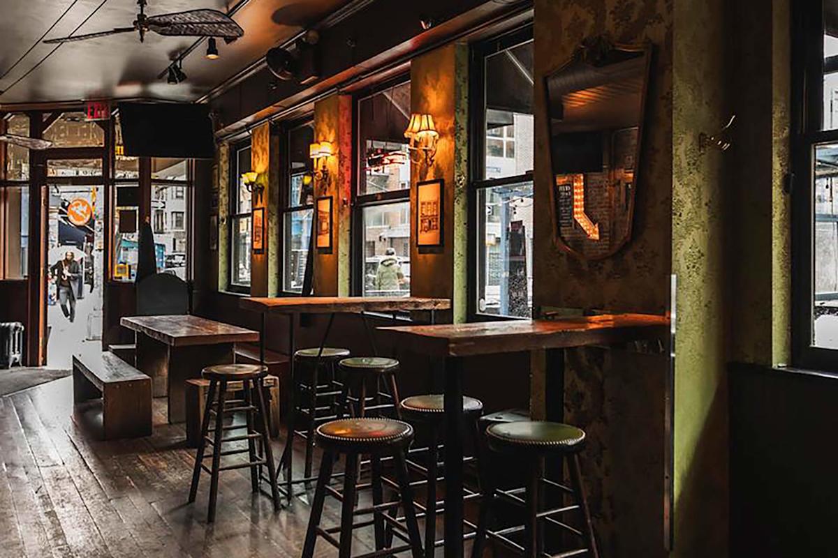 A cozy, dimly lit bar with wooden tables and stools, vintage wall lamps, a large wall mirror, and windows letting in natural light. The entrance and street are visible through the open door.
