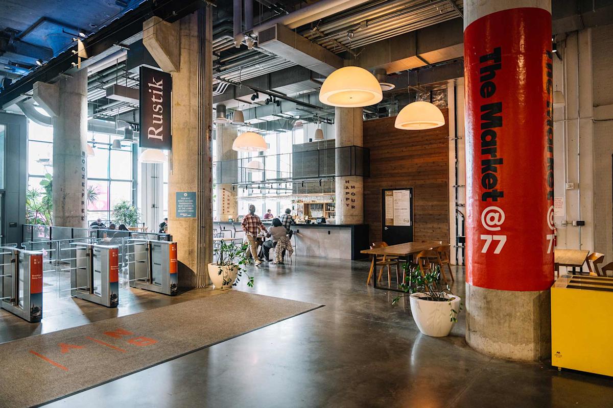 A modern, industrial-style market with high ceilings, large windows, and concrete pillars. People sit at a café-style counter. Overhead lights and indoor plants create a welcoming atmosphere. A red pillar says “The Market @ 77.”.