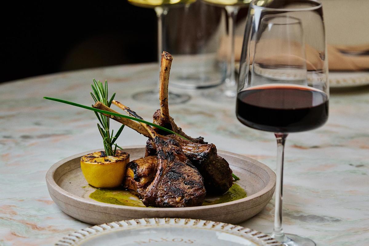 A plate of grilled lamb chops garnished with herbs and a charred lemon, served on a table next to a glass of red wine. The setting appears elegant and inviting.
