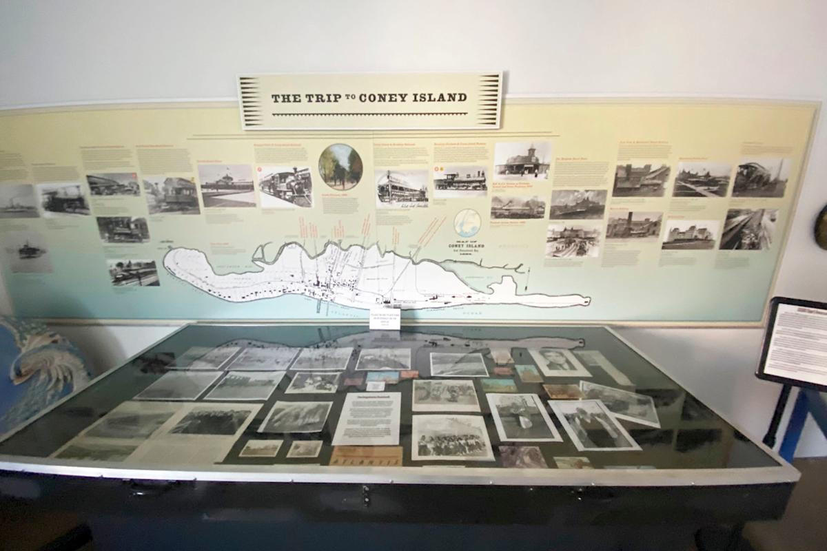 A display case features historical photos, maps, and documents related to Coney Island, with a large detailed map and informational text on the wall behind, titled "The Trip to Coney Island.