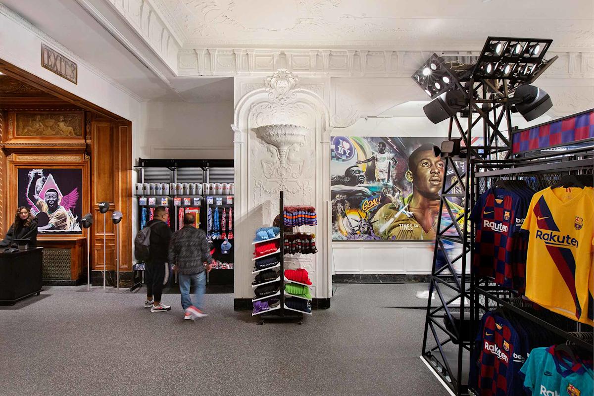 A sports merchandise store interior with racks of FC Barcelona jerseys and hats, a counter with two people, and large murals of famous athletes on ornate white walls. Two shoppers walk past the merchandise display.
