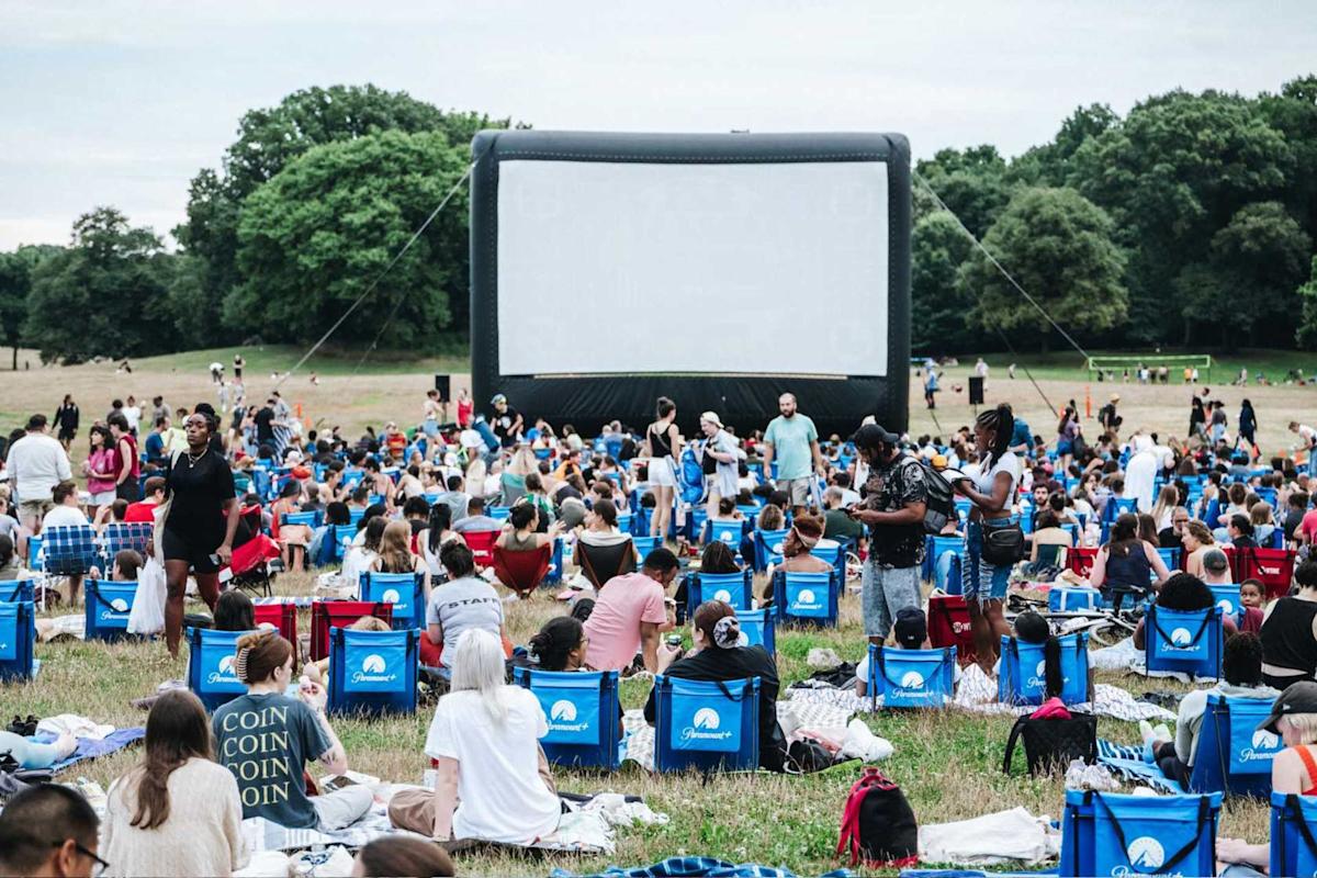 A large crowd of people sits on portable chairs and blankets in a grassy field, facing an inflatable outdoor movie screen. Trees line the background, and the atmosphere is casual and lively.