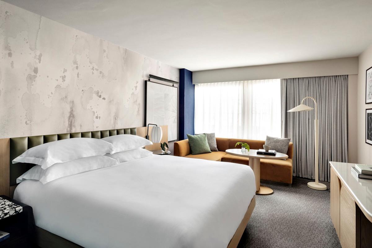 Modern hotel room with a large bed, white bedding, a tan sofa by the window, round coffee table, floor lamp, and neutral decor. Natural light streams in through a sheer-curtained window.