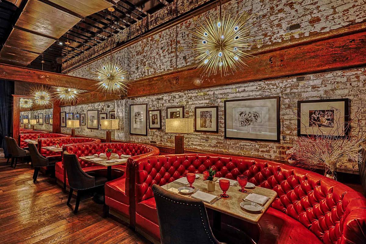 A stylish restaurant interior with red tufted leather booths, dark wood tables, elegant black chairs, and modern starburst chandeliers. Exposed brick walls display framed art and the space feels warm and inviting.
