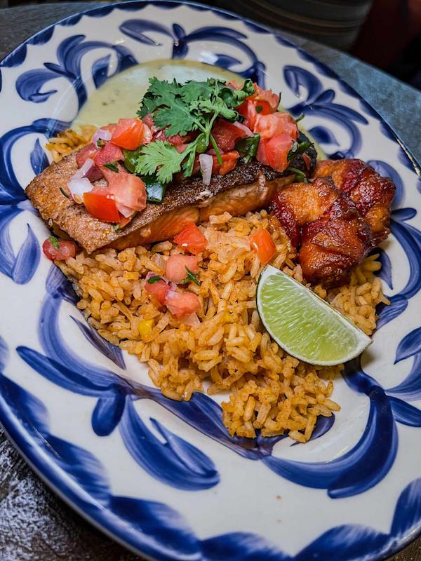 A plate with seasoned rice, a grilled fish filet topped with pico de gallo and cilantro, a piece of fried plantain, a lime wedge, and a small serving of green sauce on a decorative blue and white dish.