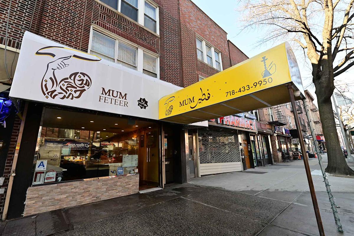 A street view of Mum Feteer, a bakery with a white and yellow awning featuring Arabic and English text, large glass windows, and a brick building exterior on a tree-lined sidewalk.