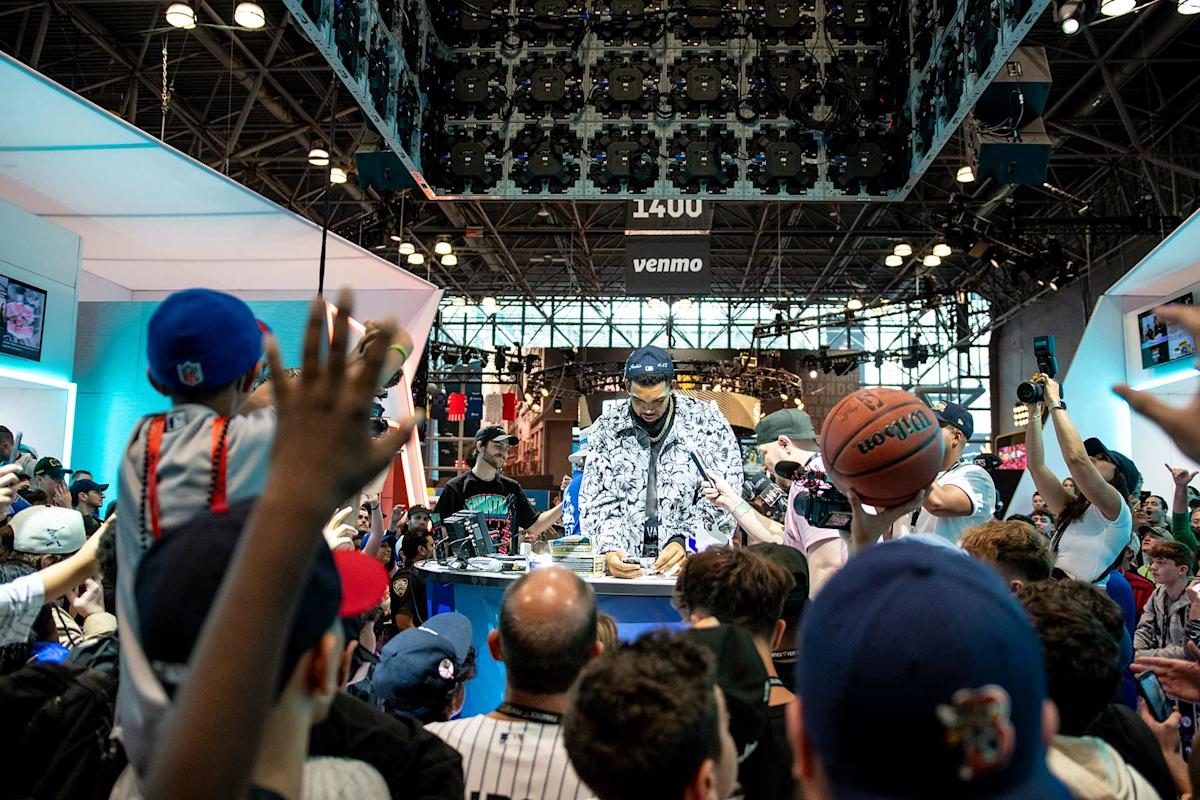 A crowd of excited fans gathers around a table where a man in patterned clothing and a cap is signing autographs at a busy indoor event; some fans hold up items like a basketball and take photos.