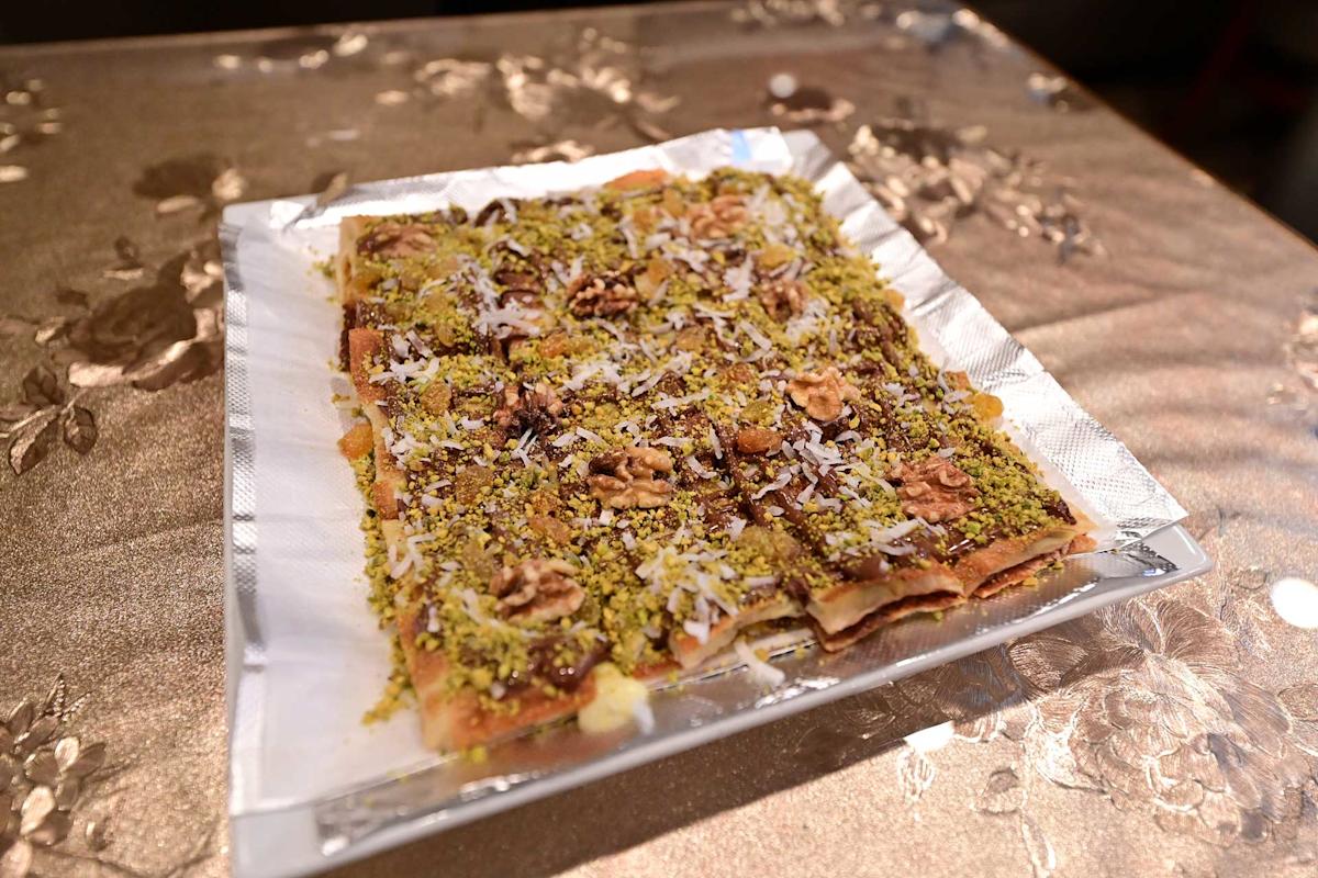 A rectangular dessert cut into square pieces, topped with chopped pistachios, walnuts, and shredded coconut, served on a white paper-lined tray on a patterned tablecloth.