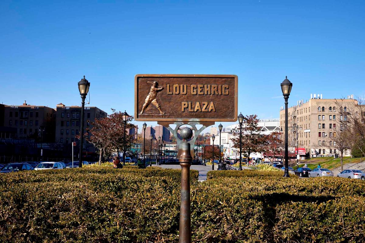 A bronze sign reading "Lou Gehrig Plaza" with a baseball player silhouette stands between two lampposts, surrounded by hedges, with buildings and parked cars in the background under a clear blue sky.