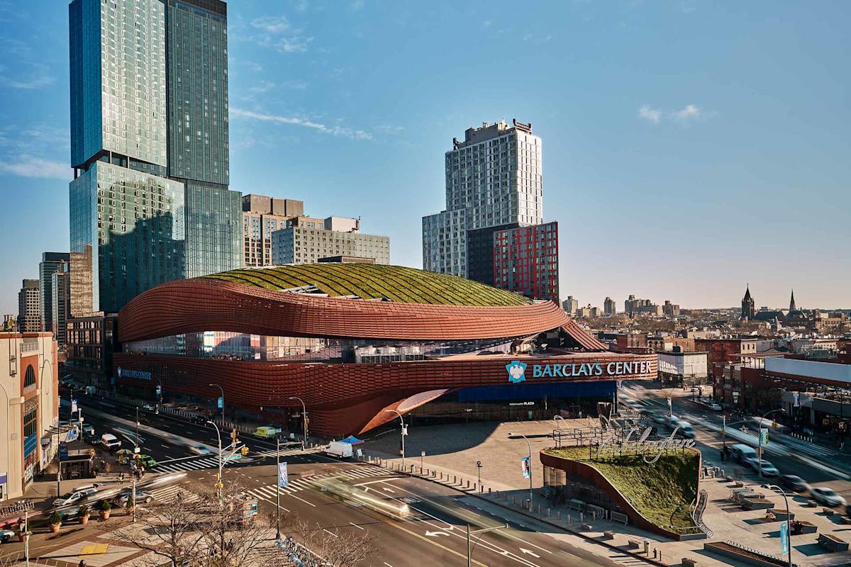 A wide view of the Barclays Center in Brooklyn, New York, showing its distinctive brown, curved design with a green roof, surrounded by city streets and tall buildings under a clear blue sky.