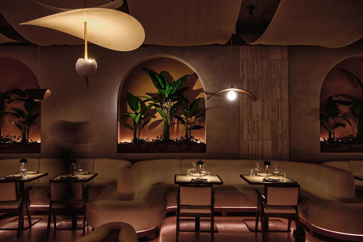 A modern, dimly lit restaurant with curved white seating, tables set for two, wall arches with green plants, and stylish pendant lighting creating a warm, intimate atmosphere.